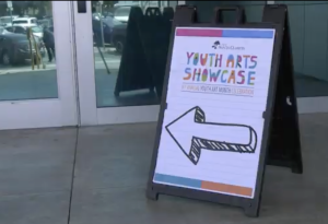 sign in front of glass doors directs visitors to the 'Youth Arts Showcase,' part of the Santa Clarita Youth Art Month Celebration. The A sign features colorful text and a large black arrow pointing to the left."